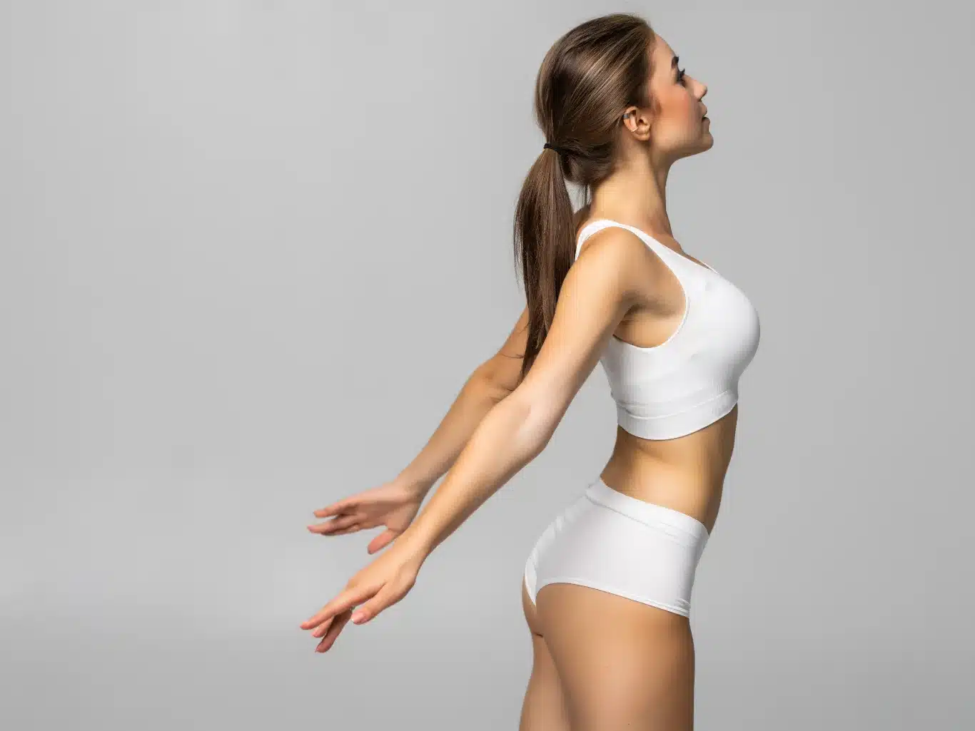 Silhouette Refinement and Body Contouring Success woman in white athletic underwear stretching her arms back showcasing a toned abdomen slim waistline and contoured glutes