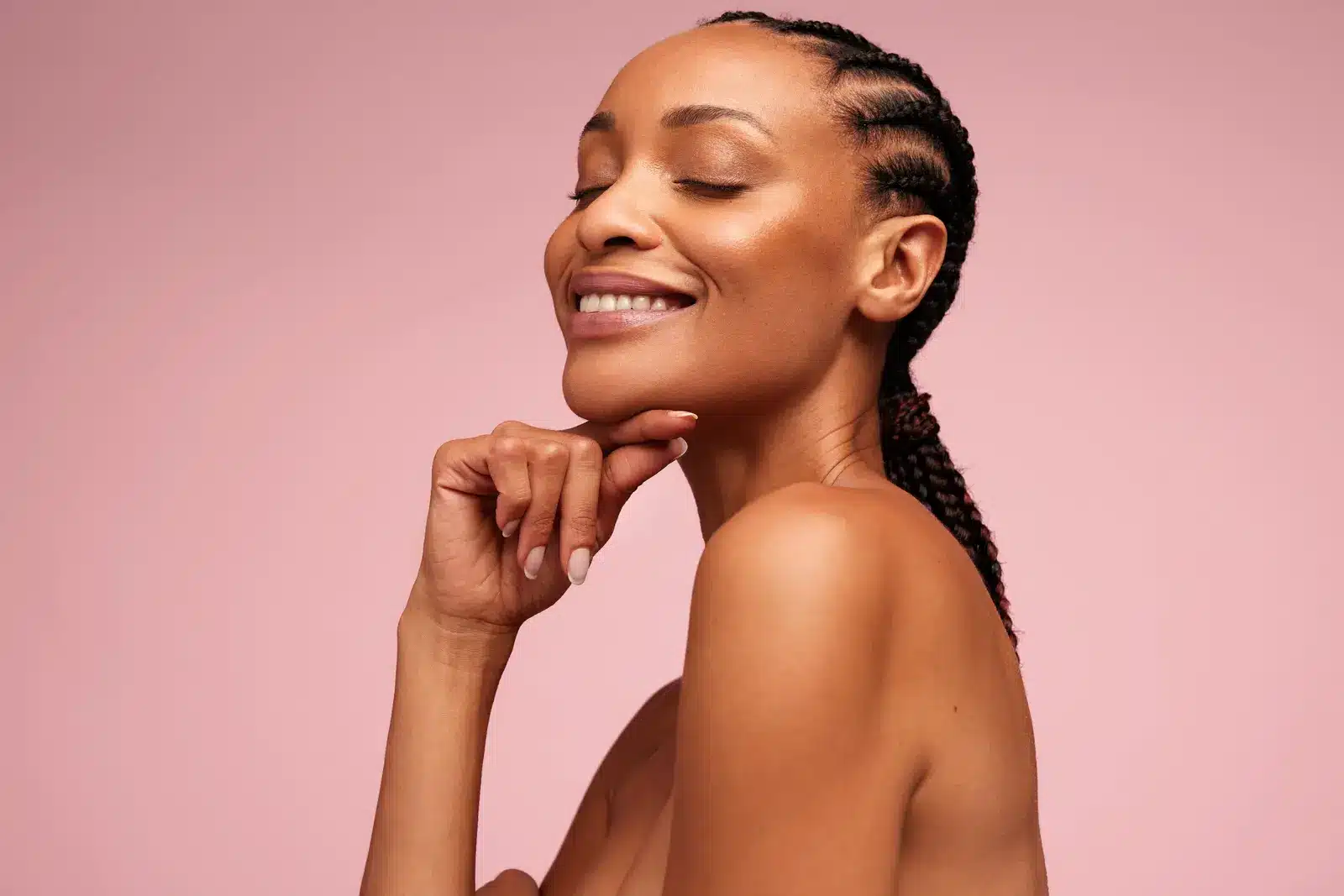 Smiling woman with braided hair and glowing skin against pink background.